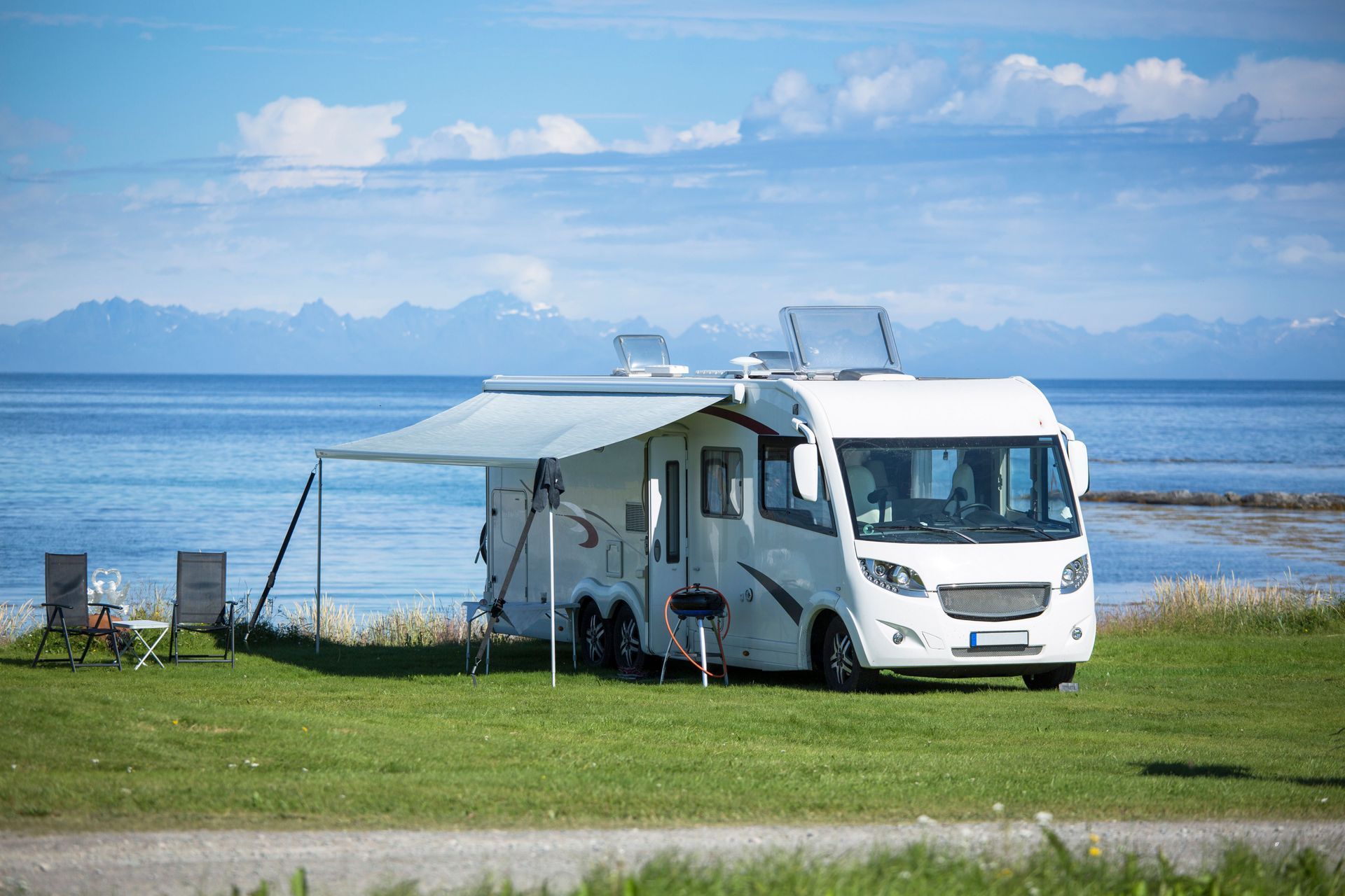 White RV parked on grassy shore, awning open, overlooking ocean and mountains; two chairs nearby.